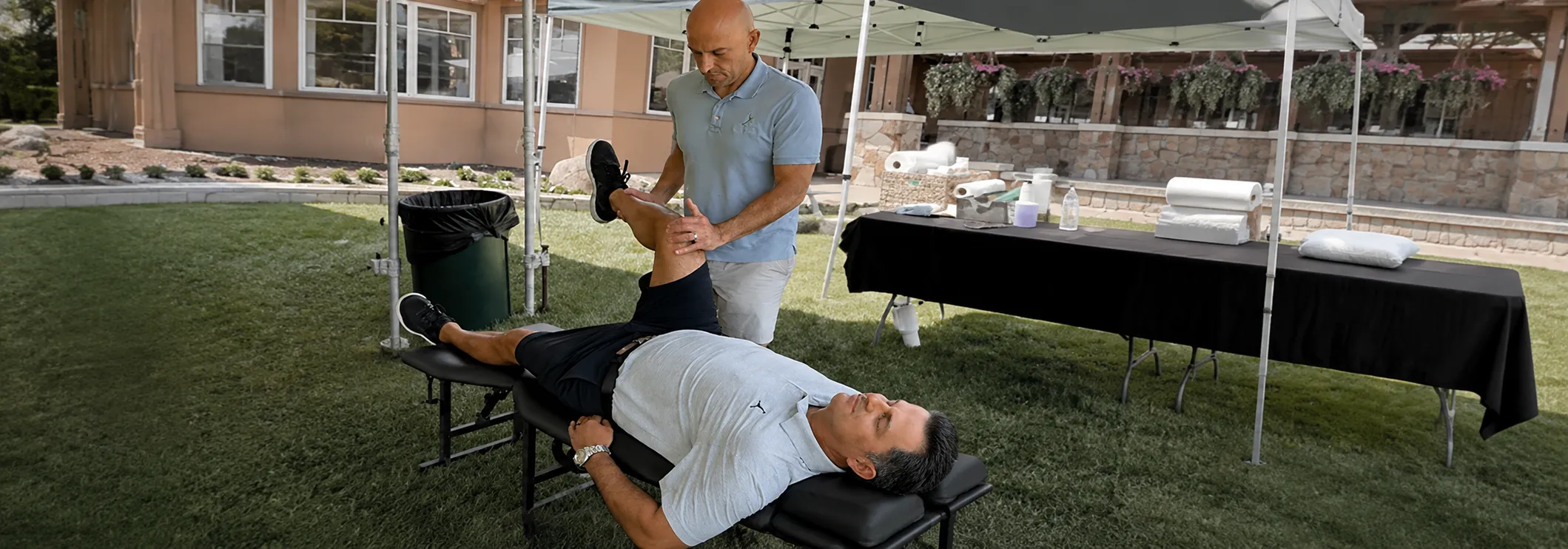 Chiropractor Armen Agacanyan guiding a patient through a leg mobility exercise on a treatment table outdoors.